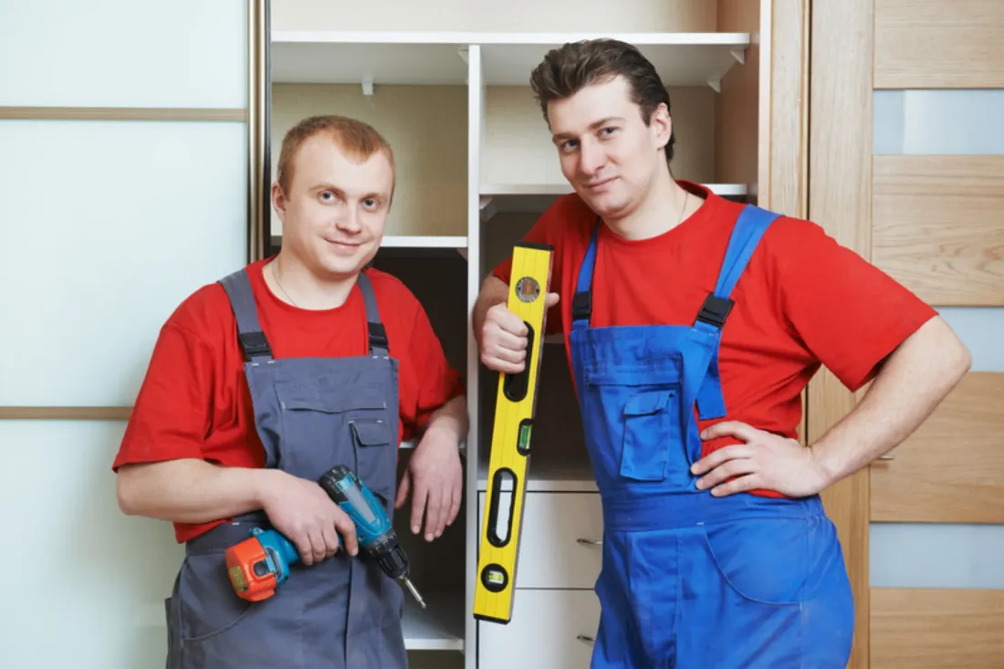 Dwóch monterów z wkrętarką i poziomicą przed montowaną szafą z białymi półkami