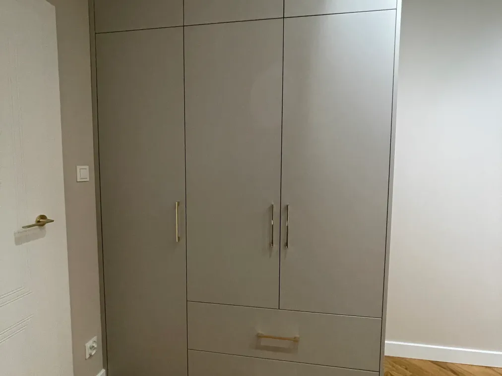 Jasnoszara szafa z trzema drzwiami, dwiema szufladami i nadstawką do sufitu, ze złotymi uchwytami, na podłodze w jodełkę